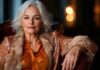 A mature woman dressed in a warm colored outfit and a stylish fur jacket sits gracefully, portraying elegance and sophistication in a thoughtfully curated space.